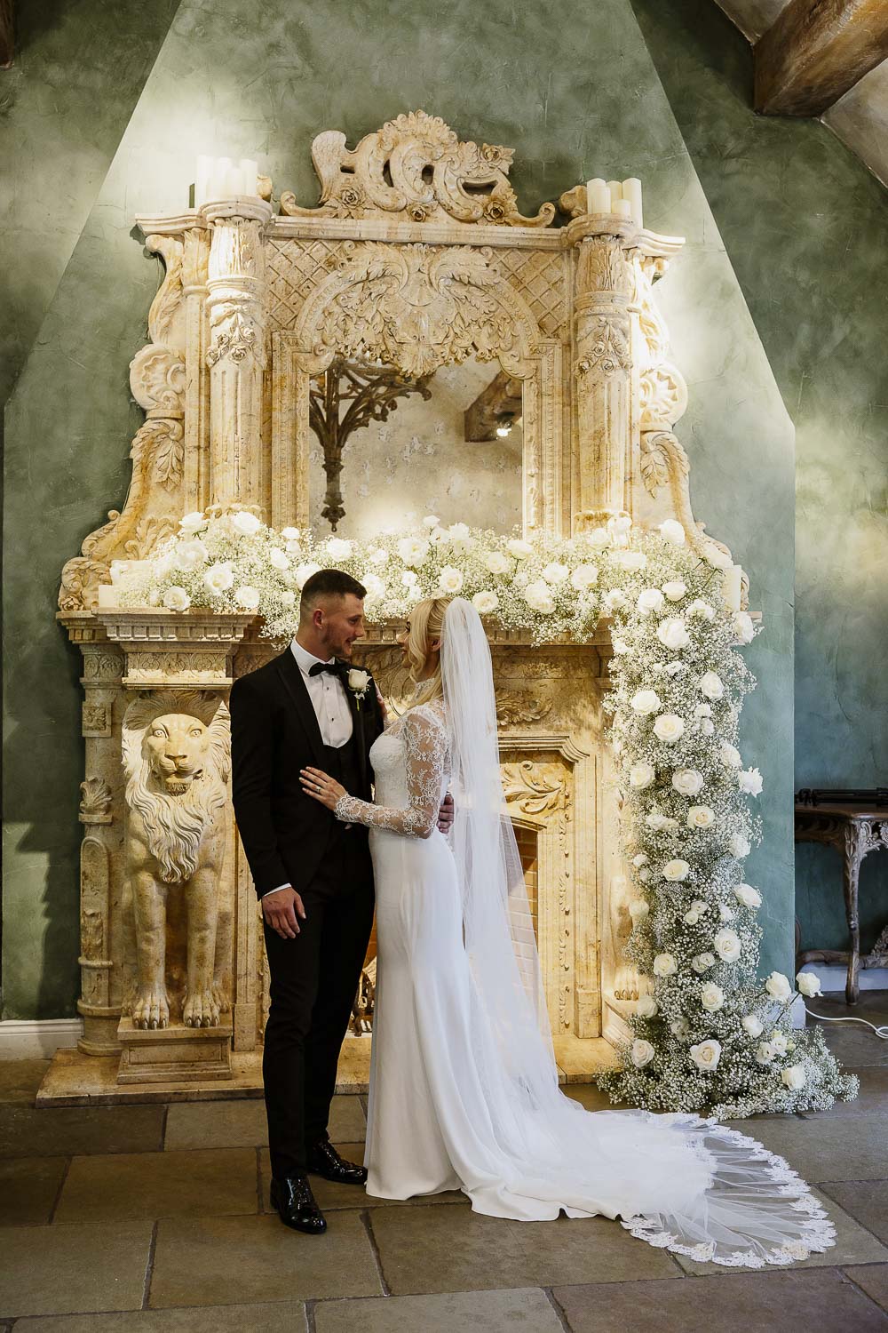 Bride and groom standing in front of the large fireplace draped in flowers at Le Petit Chateau.