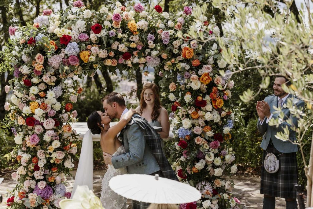 Bride and groom having their first kiss after getting married in front of an arch of fresh flowers.