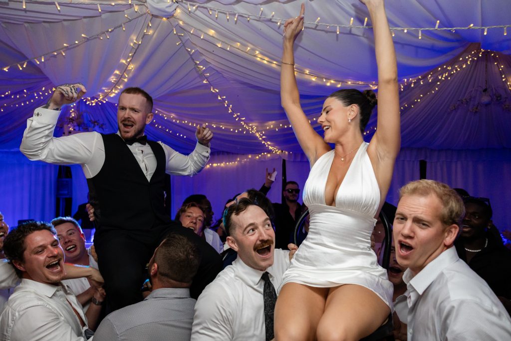Bride and groom on their wedding day, they are on the dance floor and guests have lifted the bride and groom up onto their shoulders