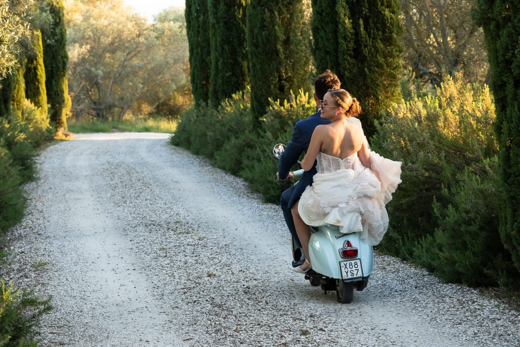 Bride and groom driving away on a vespa motorbike at Villa San Crispolto in Italy. It is their destination wedding and the bride has her wedding dress on.